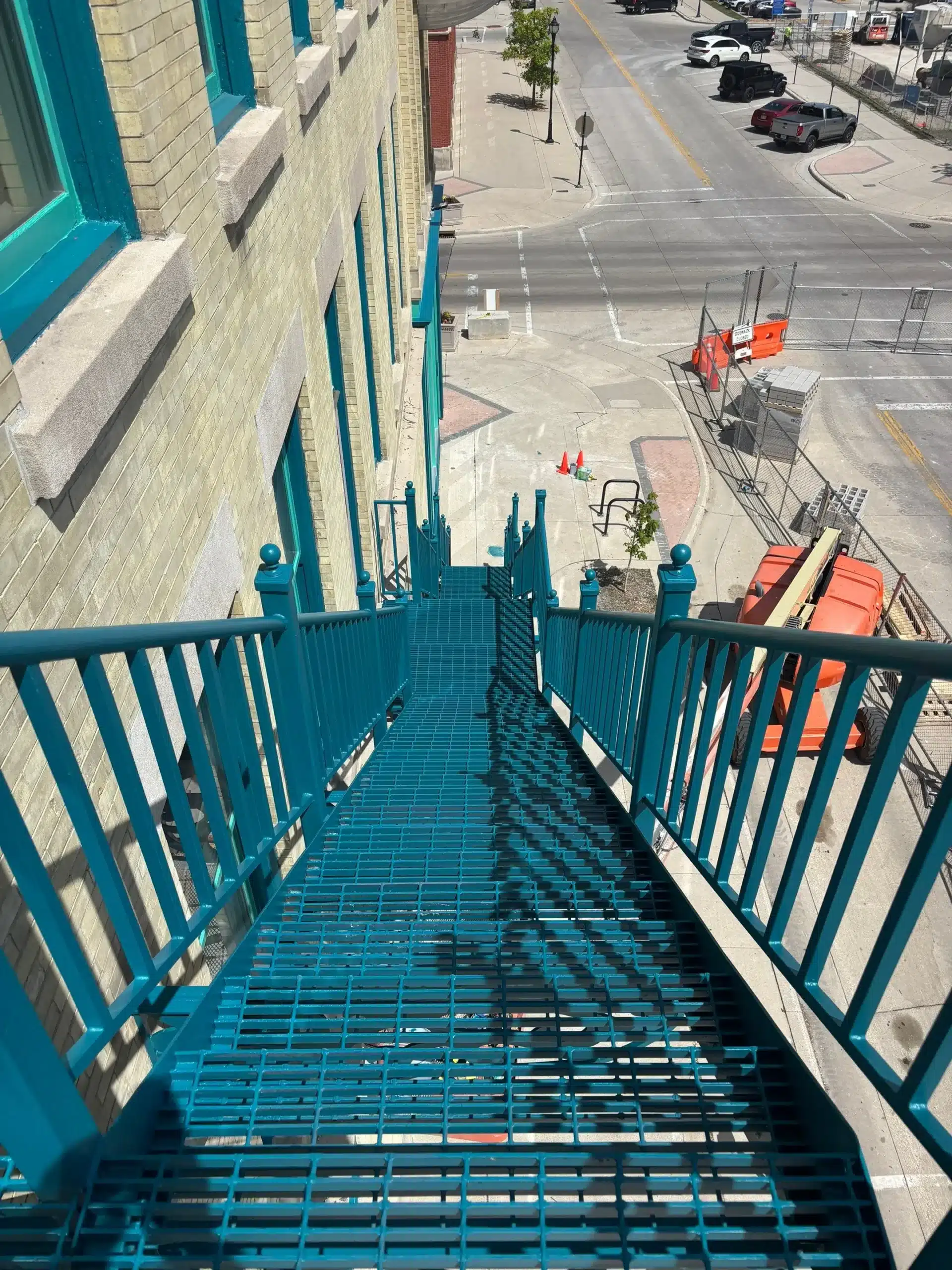 Professional Fire Escape Painting and Metal Restoration Steep teal painted metal fire escape staircase on the side of a brick commercial building, looking down to the street.